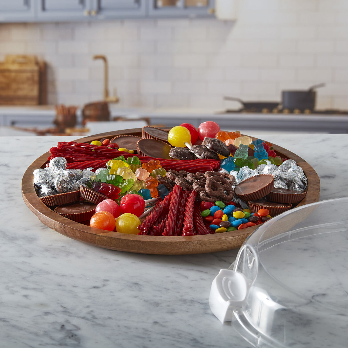 Round acacia cutting board overflowing with cheeses meats grapes berries nuts and candies on a marble kitchen counter.