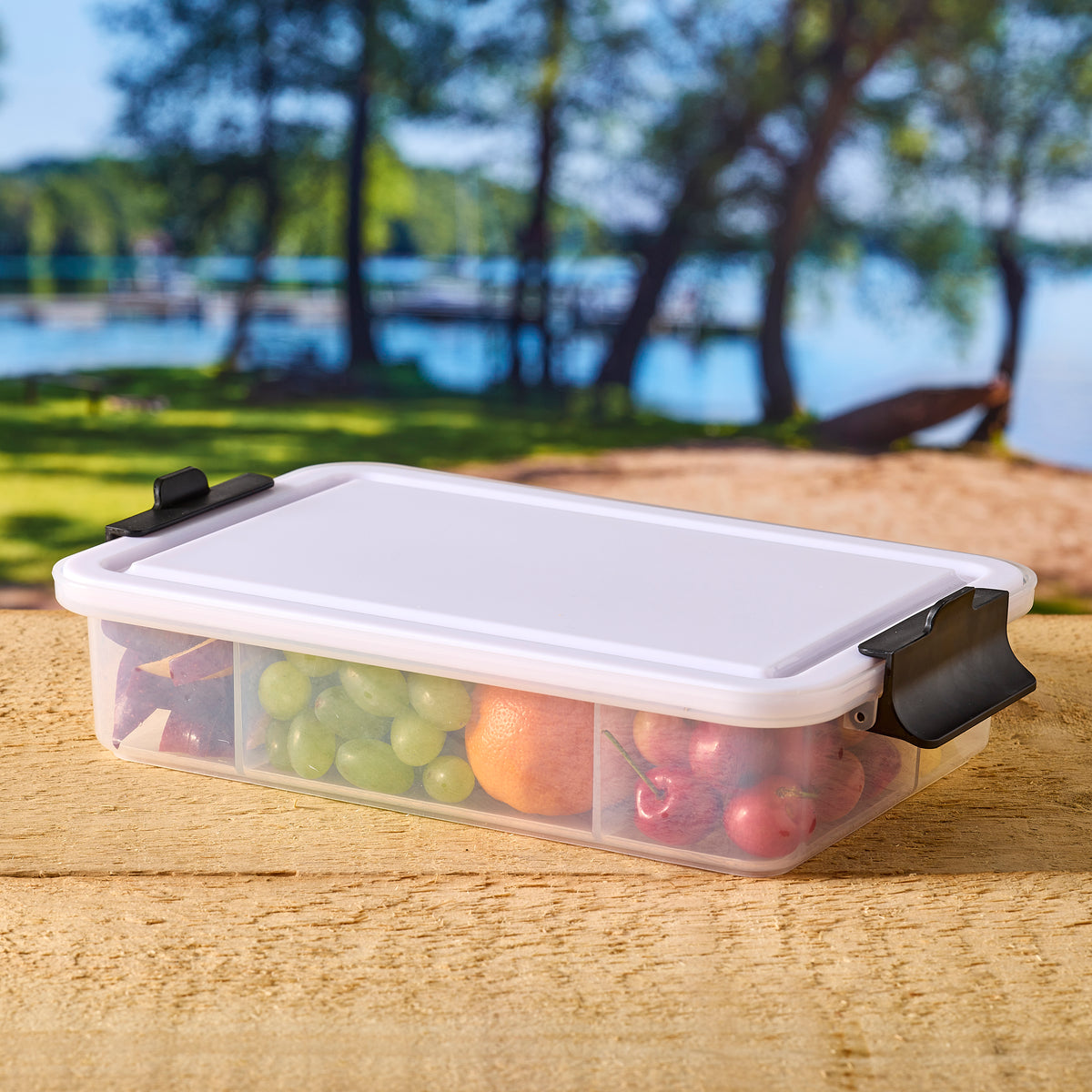 Snacklebox outdoors on a wooden surface near a lake with olives cheese and salami in the compartments
