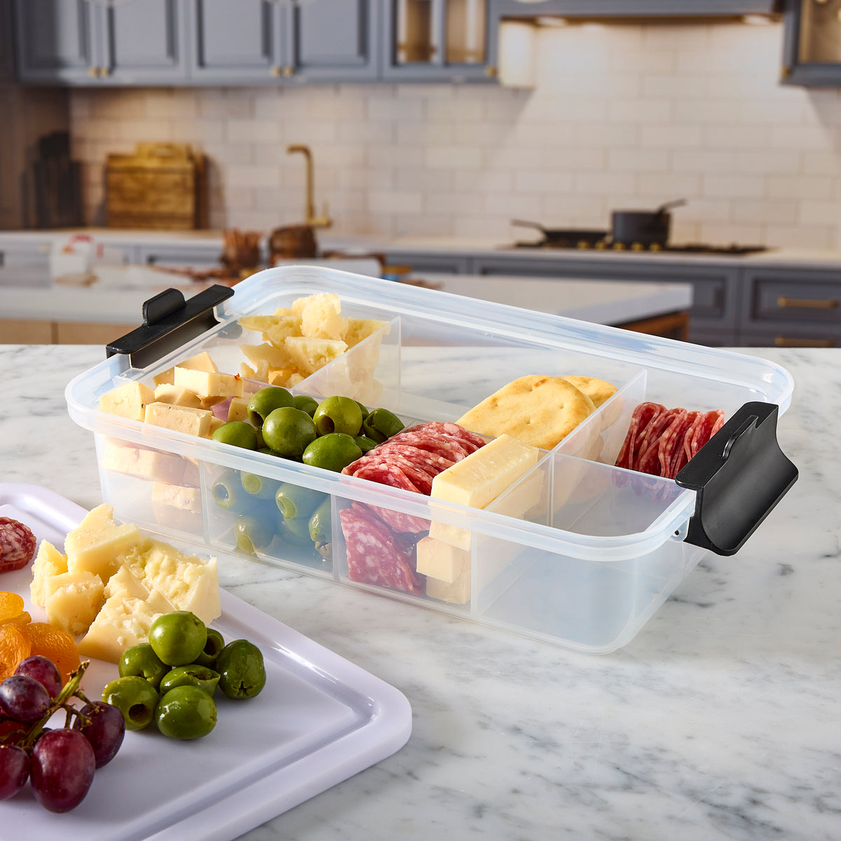 Open Snacklebox filled with cheese blocks, green olives, crackers and sliced meats on a marble kitchen counter.