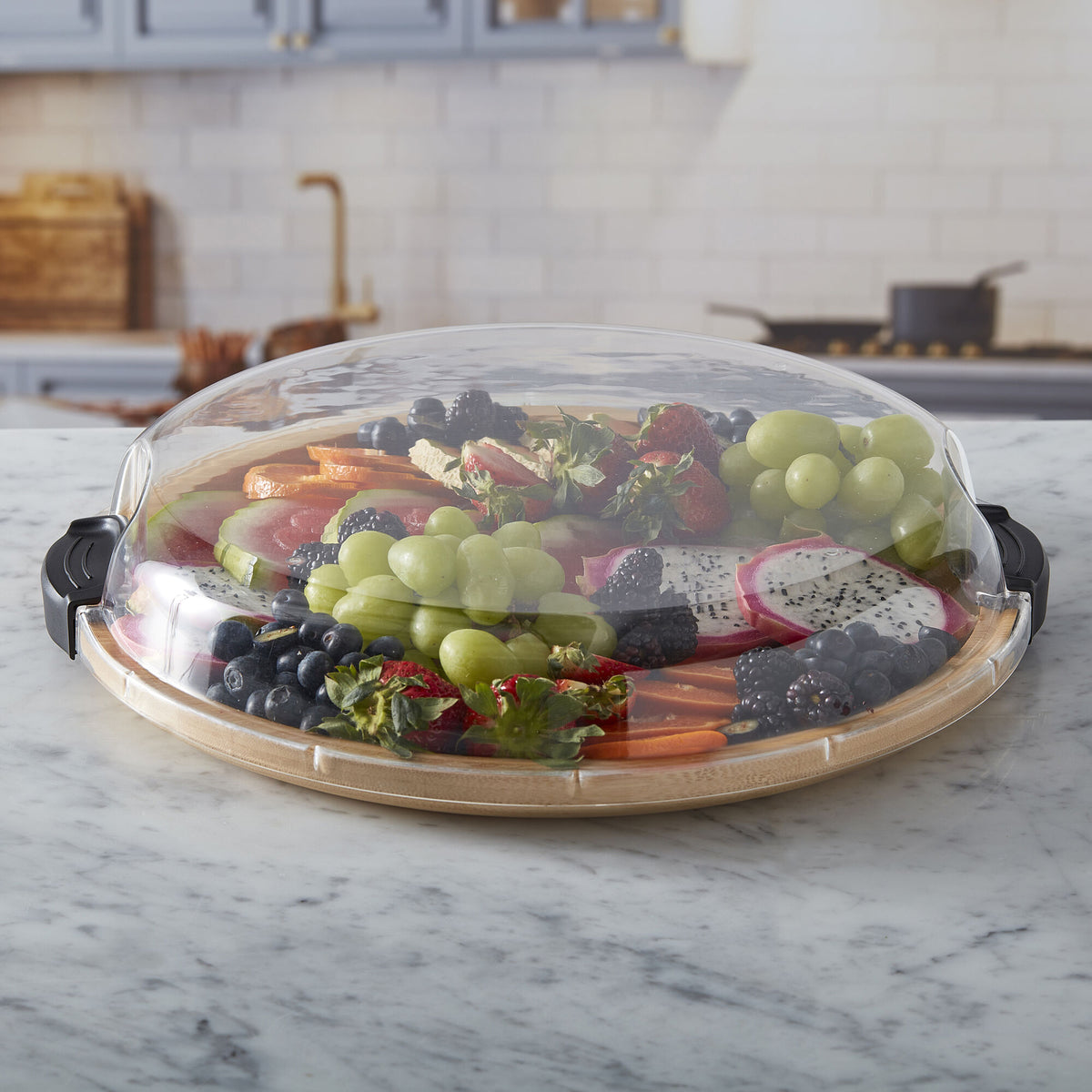 Round cutting board with a colorful fruit platter under a glass cloche on a marble countertop