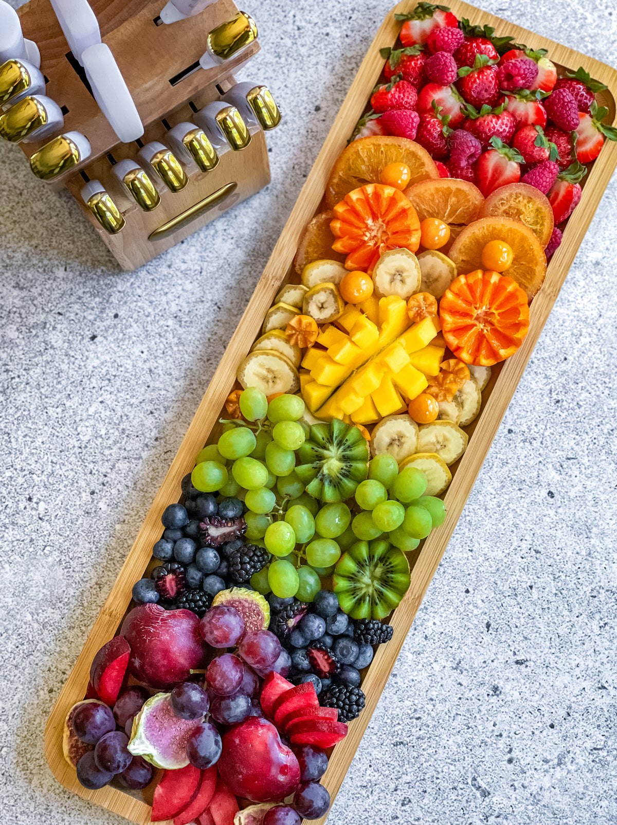 Top-down view of a Build-a-Board bamboo cutting board piled with fruit beside a knife block on a speckled countertop.