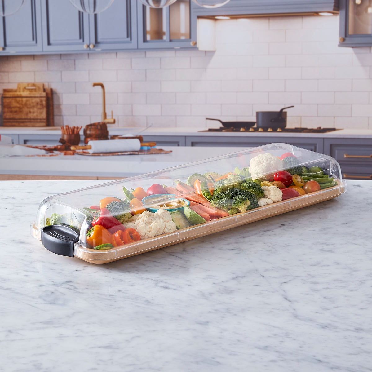 Fruit and vegetable platter on a bamboo cutting board under a clear lid on a marble counter.