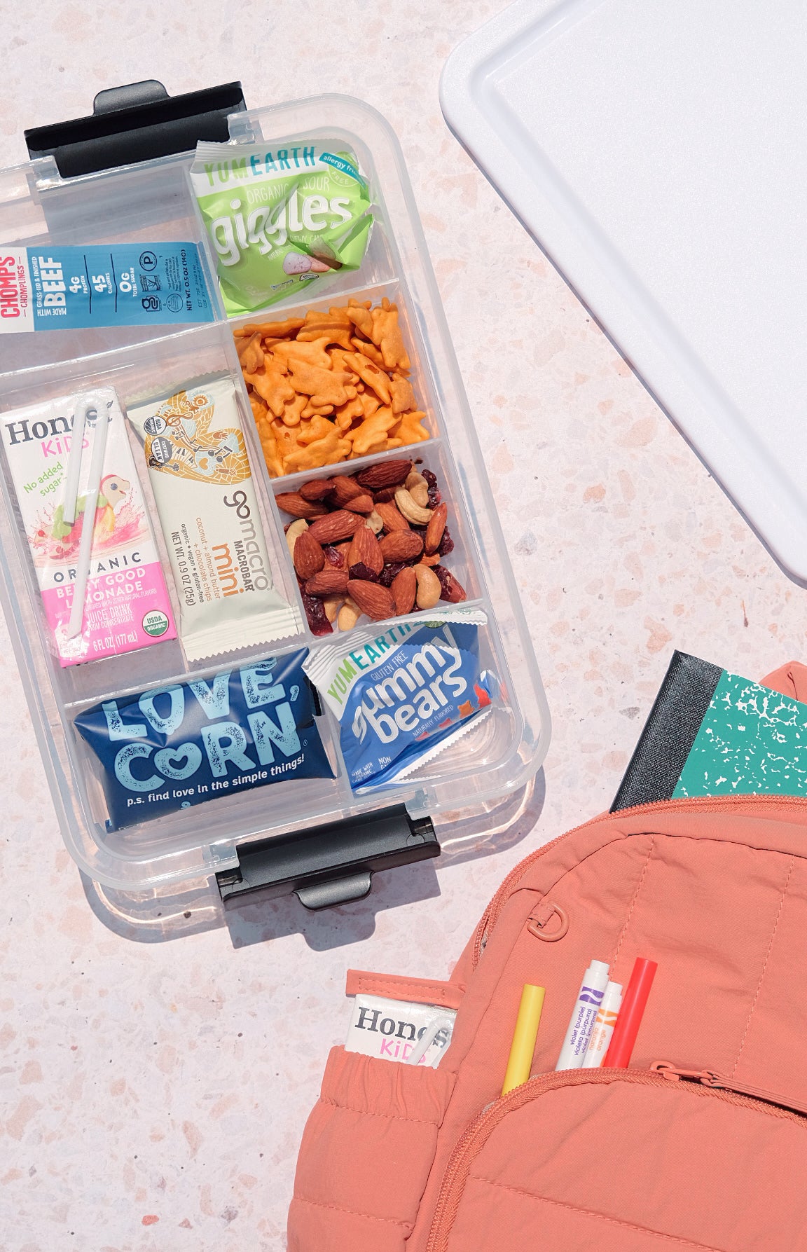 Open clear seven compartment Snacklebox filled with snacks on a kitchen counter beside a pink backpack.