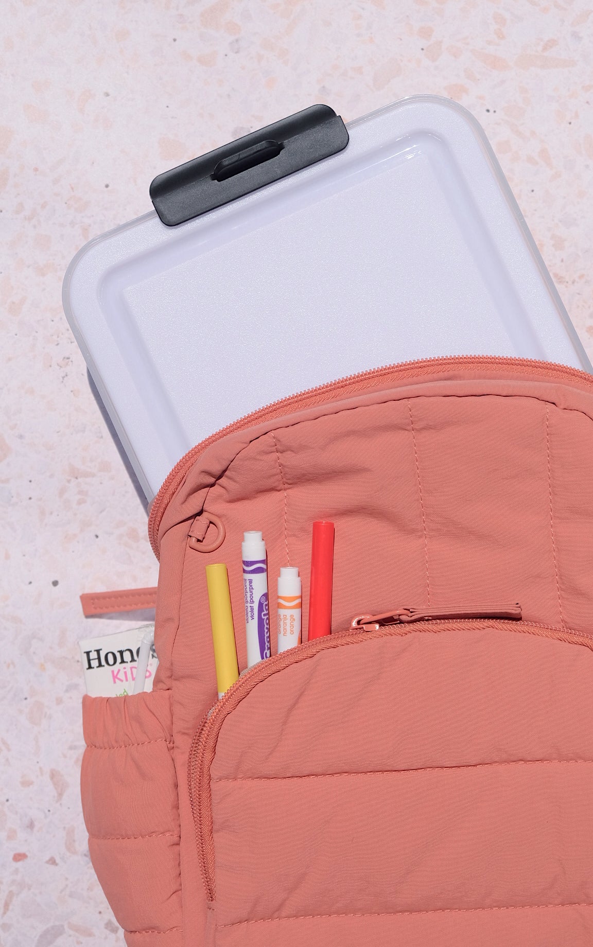 Pink backpack with markers in its side pocket beside a closed white Snacklebox on the counter.
